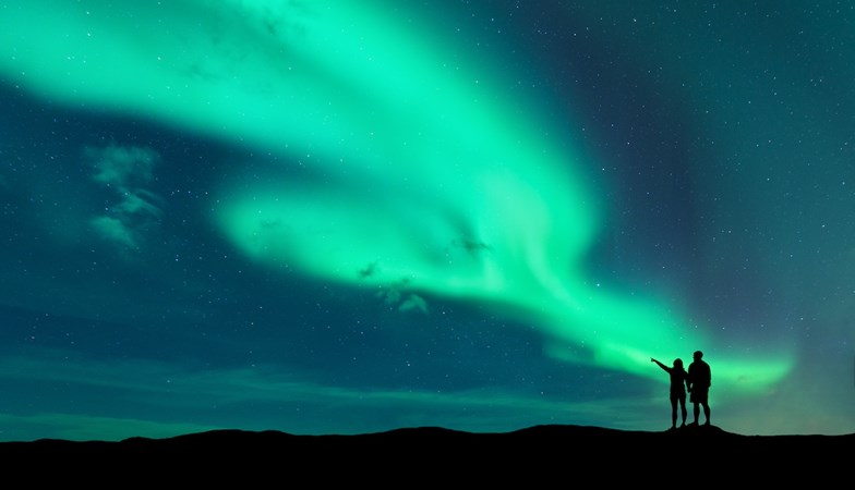 Silhuetas de duas pessoas a admirar uma aurora boreal verde vibrante num céu noturno estrelado