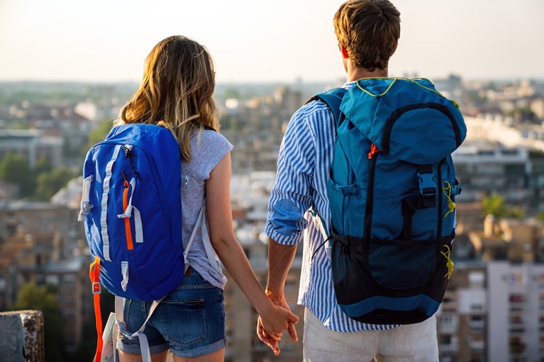 Jovem casal aventureiro, de mochila às costas, contempla a vista panorâmica de uma cidade durante uma viagem