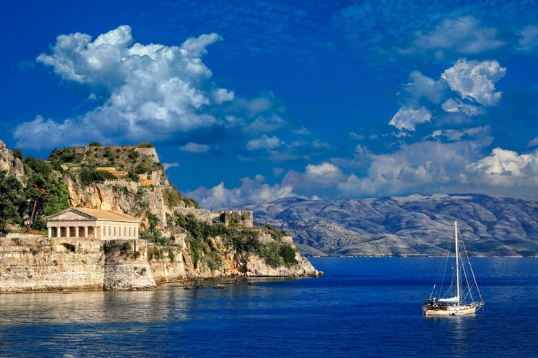 Vista da costa de Corfu com um antigo templo grego, falésias rochosas e um veleiro nas calmas águas azuis do Mar Jónico.