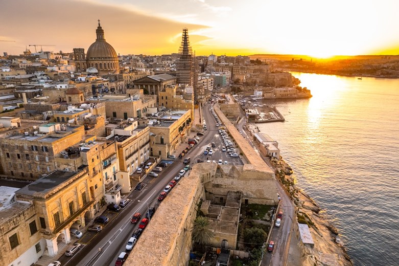 Vista panorâmica de Valletta, em Malta, ao pôr do sol, mostrando a arquitetura histórica, as ruas movimentadas e o brilho dourado sobre o porto.