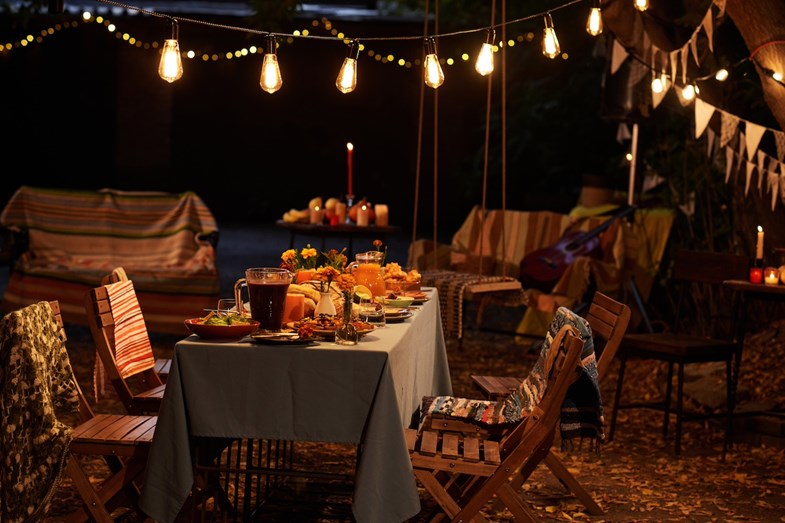 Noite. Mesa posta no jardim, luzes festivas amarelas e decoração acolhedora