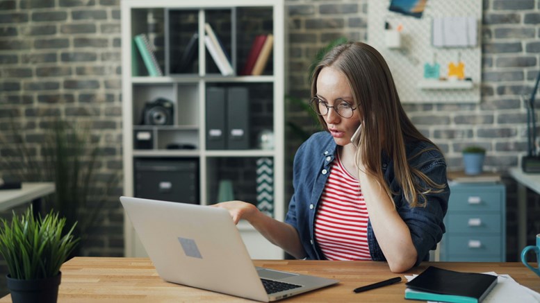 Mulher sentada numa secretária a falar ao telemóvel enquanto olha para o ecrã do computador portátil.