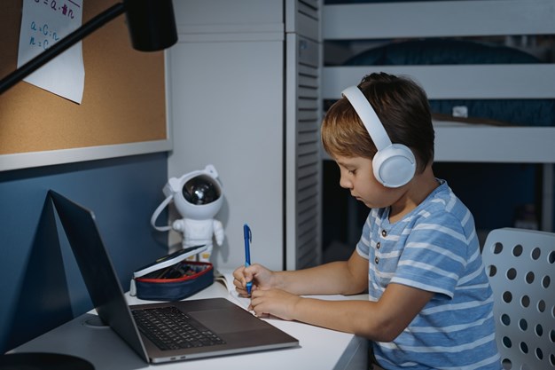 Rapaz com auscultadores e camisa às riscas, a estudar à secretária, com um computador portátil e um brinquedo robô