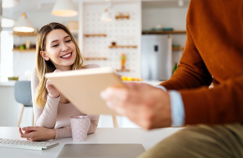 Jovem mulher sorridente na secretária observa algo que um colega mostra num tablet.