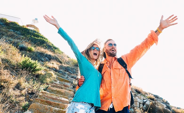 Casal sorridente abraçado, com roupas coloridas e óculos de sol, de braços levantados nas escadas do topo de uma colina