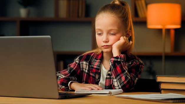 Menina sonolenta olha tenta prestar atenção a um computador portátil numa secretária ao pé de livros, numa sala com um candeeiro laranja