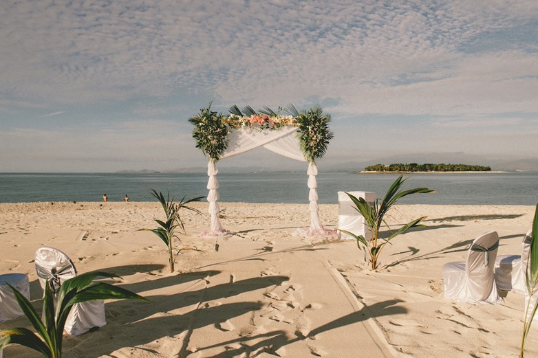 Preparativos para uma cerimónia de casamento na praia, com cadeiras brancas, decorações florais e o oceano como pano de fundo