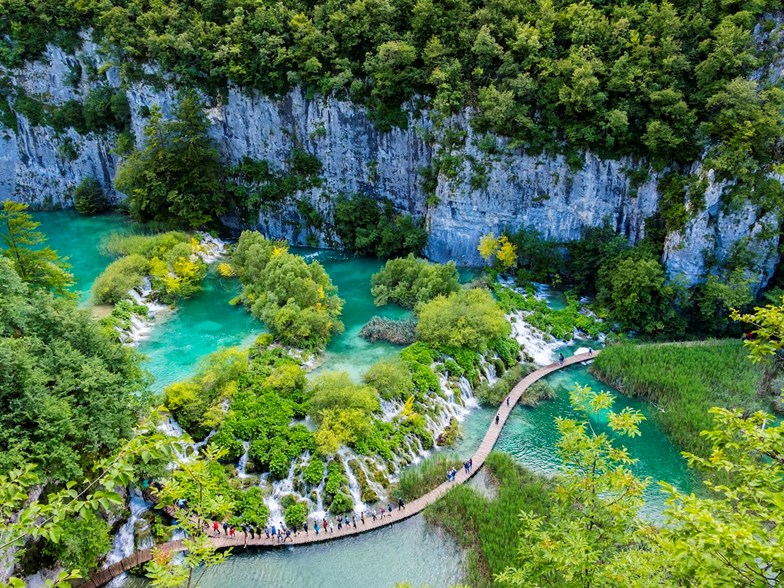 Vista aérea do Parque Nacional dos Lagos Plitvice, na Croácia, com lagos azul-turquesa, cascatas e um passadiço de madeira com turistas.