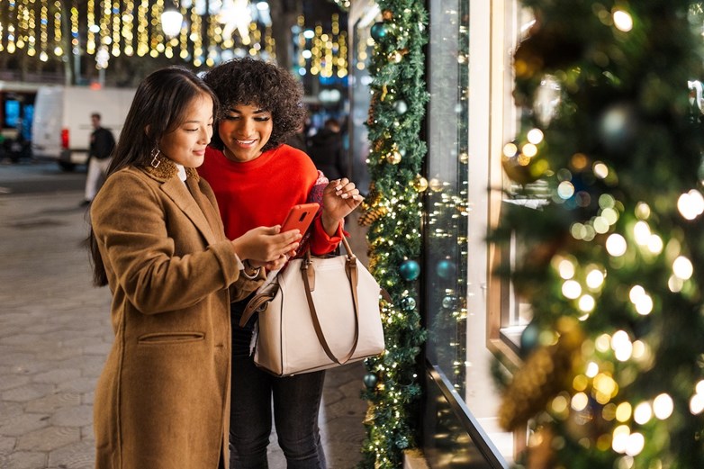 Duas mulheres a olhar para o telemóvel enquanto fazem compras a aproveitar os descontos da Black Friday numa rua decorada com luzes de Natal.