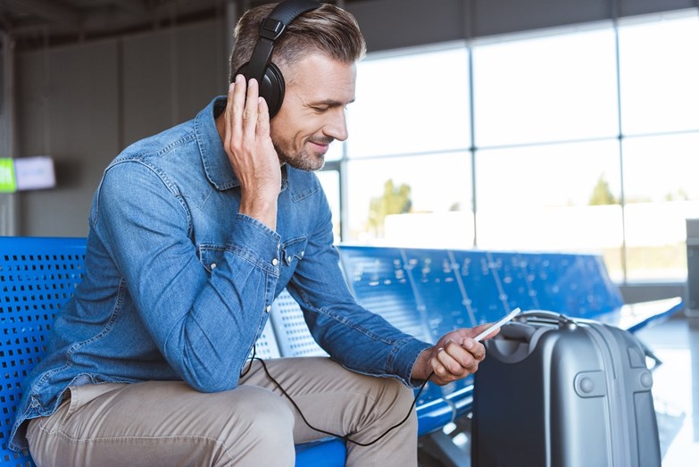 Homem com auscultadores olha para o smartphone enquanto está sentado com a bagagem no aeroporto.